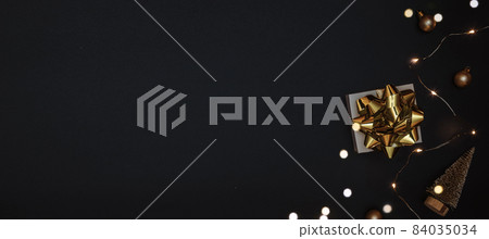 Christmas gift. White gift with golden bow, gold balls and sparkling lights garland in xmas decoration on dark background for greeting card. Xmas backdrop with space for text. 84035034