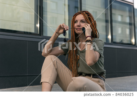 Young woman with red dreadlocks thoughtfully looks into the distance. Talking on the phone. Street photo 84036025