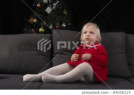 Little blonde girl in a red dress sits on the sofa against the background of a Christmas tree and a black wall. Christmas Eve. New Year 84036125