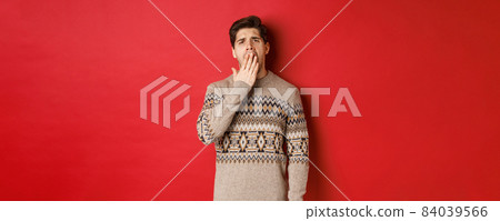 Image of tired or bored handsome man in winter sweater, yawning and covering mouth with hand, standing exhausted against red background 84039566