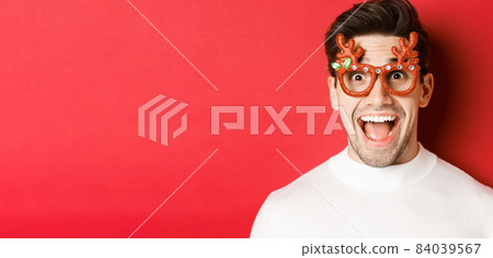 Concept of winter holidays, christmas and celebration. Close-up of attractive guy with bristle, wearing new year party glasses and looking amazed at promo offer, standing over red background 84039567