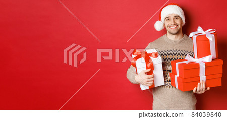Concept of winter holidays, new year and celebration. Portrait of lovely smiling man prepared gifts for christmas, holding presents and looking at camera heartwarming, red background Concept of winter holidays, new year and celebration. Portrait of lovely smiling man prepared gifts for christmas, holding presents and looking at camera heartwarming, red background 84039840