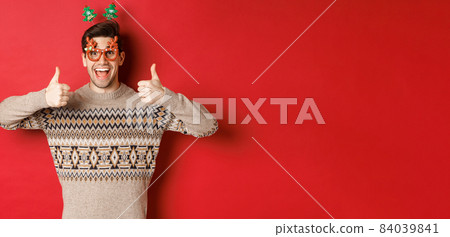 Portrait of satisfied and happy man in christmas sweater and party glasses, showing thumbs-up, wishing happy new year, standing over red background Portrait of satisfied and happy man in christmas sweater and party glasses, showing thumbs-up, wishing happy new year, standing over red background 84039841