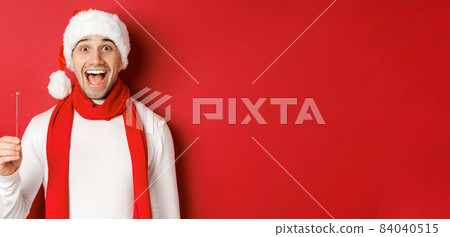 Concept of christmas, winter holidays and celebration. Close-up of happy handsome man in santa hat and scarf, having fun on new year party, holding sparkler and smiling, red background 84040515