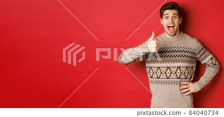 Portrait of excited, attractive man in christmas sweater, showing thumbs-up and looking amazed, recommending good thing, advertising christmas related product Portrait of excited, attractive man in christmas sweater, showing thumbs-up and looking amazed, recommending good thing, advertising christmas related product 84040734