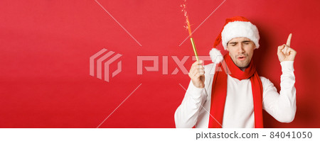 Concept of christmas, winter holidays and celebration. Attractive guy enjoying new year party, dancing with sparkler, wearing santa hat and scarf, standing over red background 84041050