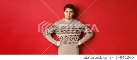 Portrait of doubtful and disappointed man, wearing christmas sweater, frowning and looking displeased at you, standing against red background Portrait of doubtful and disappointed man, wearing christmas sweater, frowning and looking displeased at you, standing against red background 84041200