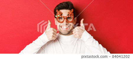 Concept of winter holidays, christmas and celebration. Close-up of excited handsome man in party glasses, smiling and showing thumbs-up in approval, standing over red background Concept of winter holidays, christmas and celebration. Close-up of excited handsome man in party glasses, smiling and showing thumbs-up in approval, standing over red background 84041202
