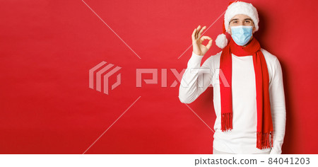 Concept of covid-19, christmas and holidays during pandemic. Portrait of happy and satisfied man in medical mask and santa hat, showing okay sign in approval, recommending something 84041203