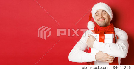 Concept of winter holidays, christmas and lifestyle. Image of lovely guy in santa hat and scarf, hugging his new year present and smiling flattered, standing over red background Concept of winter holidays, christmas and lifestyle. Image of lovely guy in santa hat and scarf, hugging his new year present and smiling flattered, standing over red background 84041206