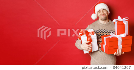 Concept of winter holidays, new year and celebration. Portrait of lovely smiling man receiving pile of presents, holding gifts and being touched with surprise, standing over red background grateful 84041207