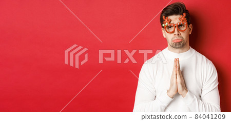 Image of handsome man in christmas party glasses, begging for help or apologizing, need a favour, standing over red background Image of handsome man in christmas party glasses, begging for help or apologizing, need a favour, standing over red background 84041209