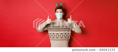 Concept of new year, covid-19 and social distancing. Cheerful caucasian man in sweater and medical mask, celebrating christmas during pandemic, showing thumbs-up, red background Concept of new year, covid-19 and social distancing. Cheerful caucasian man in sweater and medical mask, celebrating christmas during pandemic, showing thumbs-up, red background 84041333