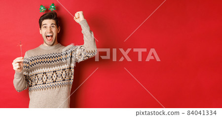 Cheerful guy with a sparkler rejoicing and wishing happy new year, raising hand up and shouting for joy, standing over red background Cheerful guy with a sparkler rejoicing and wishing happy new year, raising hand up and shouting for joy, standing over red background 84041334