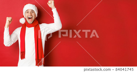 Image of joyful caucasian man in santa hat and scarf, shouting for joy and raising hands, celebrating victory or win, triumphing over red background 84041745