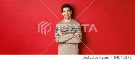 Portrait of cheerful, attractive man in christmas sweater, laughing and smiling, celebrating new year and winter holidays, standing over red background 84041875