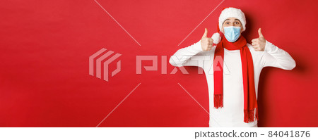 Concept of new year, coronavirus and holidays. Cheerful man celebrating new year and social distancing, wearing medical mask, santa hat and scarf, showing thumbs-up in approval 84041876