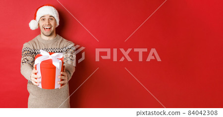 Image of happy man saying merry christmas and giving you a gift in box, smiling cheerful, celebrating winter holidays in santa hat and xmas sweater, standing over red background Image of happy man saying merry christmas and giving you a gift in box, smiling cheerful, celebrating winter holidays in santa hat and xmas sweater, standing over red background 84042308