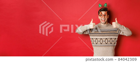 Portrait of satisfied and happy man in christmas sweater and party glasses, showing thumbs-up, wishing happy new year, standing over red background 84042309