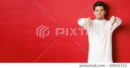 Portrait of disappointed skeptical guy, wearing white sweater, showing thumbs-down to disagree, express dislike, standing over red background Portrait of disappointed skeptical guy, wearing white sweater, showing thumbs-down to disagree, express dislike, standing over red background 84042312