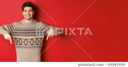 Concept of christmas celebration, winter holidays and lifestyle. Attractive happy man in xmas sweater showing promotion, pointing fingers down at logo, standing over red background 84042484