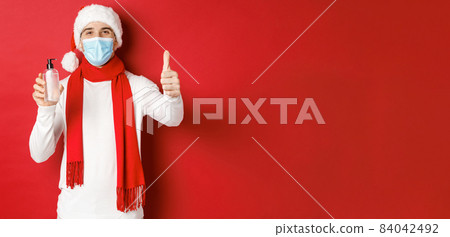 Concept of covid-19, christmas and holidays during pandemic. Happy and satisfied man in santa hat and medical mask, showing thumb-up in approval and recommending hand sanitizer Concept of covid-19, christmas and holidays during pandemic. Happy and satisfied man in santa hat and medical mask, showing thumb-up in approval and recommending hand sanitizer 84042492