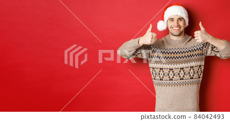 Portrait of attractive happy man in santa hat and sweater, showing thumbs-up in approval and smiling, wishing merry christmas, standing over red background Portrait of attractive happy man in santa hat and sweater, showing thumbs-up in approval and smiling, wishing merry christmas, standing over red background 84042493