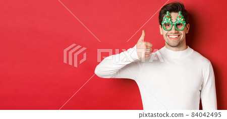 Concept of christmas, winter holidays and celebration. Close-up of handsome smiling man, wearing party glasses and white sweater, showing thumbs-up, recommending new year promo 84042495