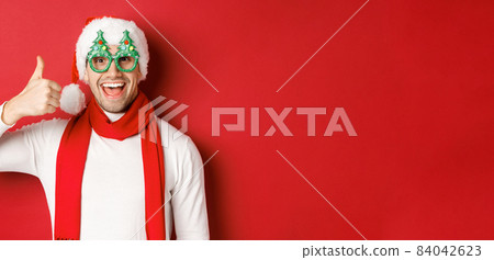 Concept of christmas, winter holidays and celebration. Cheerful guy in santa hat and party glasses, showing thumb-up and smiling pleased, standing over red background Concept of christmas, winter holidays and celebration. Cheerful guy in santa hat and party glasses, showing thumb-up and smiling pleased, standing over red background 84042623