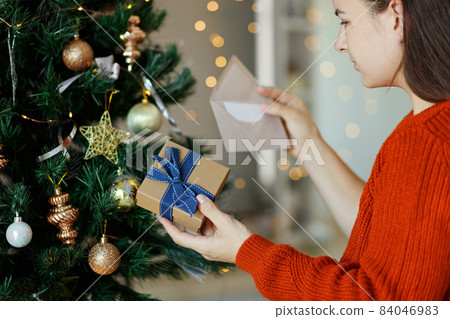 Woman opening gift box next to xmas tree and reading greeting card. Christmas time, New Year holidays. 84046983