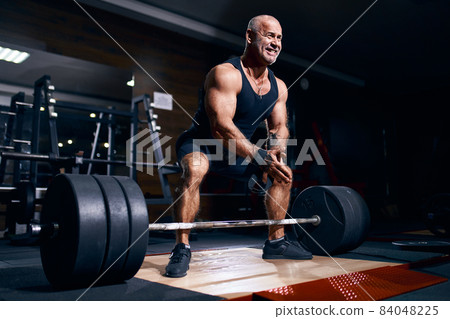 Older bodybuilder preparing to exercise deadlift with barbell while on cross training in a gym. 84048225