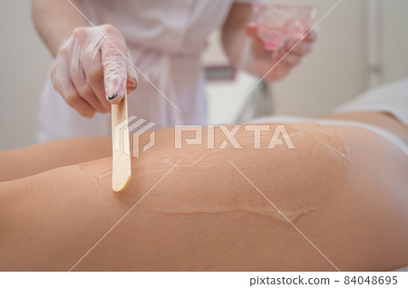The master applies the anesthetic gel with a spatula to the woman's legs before laser hair removal. Permanent hair removal with a device in the clinic The master applies the anesthetic gel with a spatula to the woman's legs before laser hair removal. Permanent hair removal with a device in the clinic 84048695