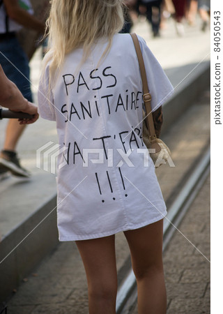 portrait on back view of woman protesting in the street with text on shirt in french, pass sanitaire vat faire !!!, in English, sanitary pass F... you !!! portrait on back view of woman protesting in the street with text on shirt in french, pass sanitaire vat faire !!!, in English, sanitary pass F... you !!! 84050543