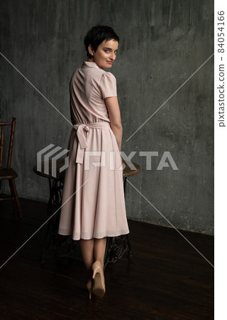 Young brunette woman in long old-fashioned pink dress stands near table turning around for posing on camera Young brunette woman in long old-fashioned pink dress stands near table turning around for posing on camera 84054166