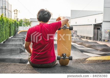 Asian man sitting with surfskate or skate board in pumptrack skate Park when sunrise time over photo blur of pumptrack curve, extream sport, healthy and exercise, fashion in covid19 concept Asian man sitting with surfskate or skate board in pumptrack skate Park when sunrise time over photo blur of pumptrack curve, extream sport, healthy and exercise, fashion in covid19 concept 84056360