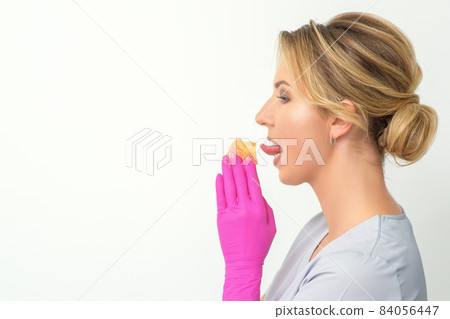 Young beautician licks liquid wax for depilation holding in gloved hands standing on white background. Cosmetologist tastes the sugar paste to taste. 84056447