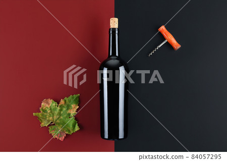 Top view of bottle of red wine with green fall grapes leaf and corkscrew on red and black geometric background 84057295