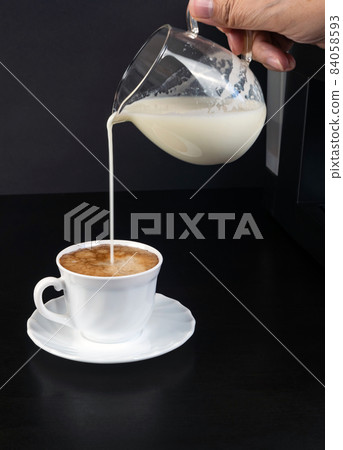 Milk is pouring from a jug into a white cup of coffee with froth on a black background. Vertical photo Milk is pouring from a jug into a white cup of coffee with froth on a black background. Vertical photo 84058593