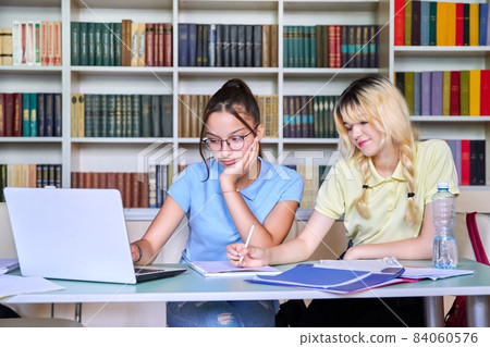 Two teenage schoolgirls students sitting with a laptop in the library Two teenage schoolgirls students sitting with a laptop in the library 84060576