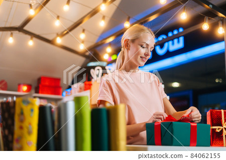 Low-angle view of young smiling woman designer wrapping Christmas gift box tying red ribbon and decorating beautiful bow standing at table of holiday store 84062155
