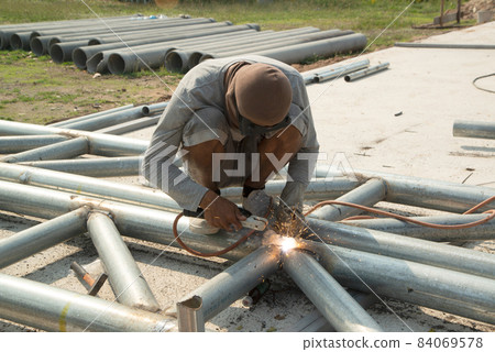 An Asian welder is sitting on the steel roof frame and is welding. 84069578