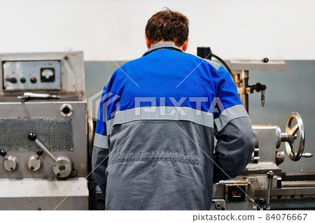 Turner stands behind lathe in production hall and works. View of worker from behind from back in 84076667