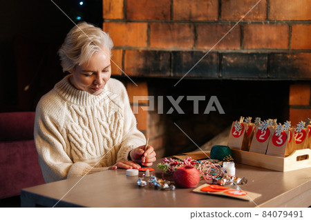 Happy young woman writing number on red bag making paper bags from kraft paper for advent calendar on Christmas Eve sitting by fireplace. 84079491