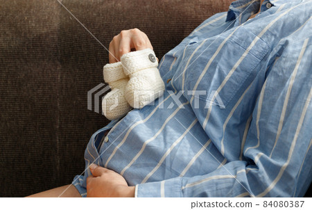 A pregnant woman in a blue shirt, holding knitted white shoes on her tummy 84080387