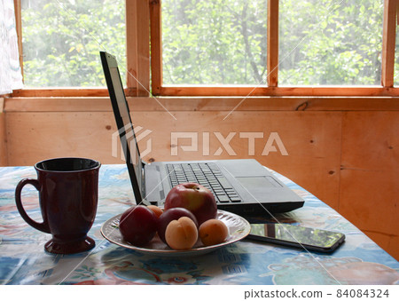 In a country house, there is a laptop, a plate of fruit, a smartphone on the table. Remote work concept, online training 84084324