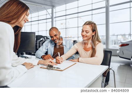 Beautiful young couple signs documents at car dealership showroom Beautiful young couple signs documents at car dealership showroom 84084809