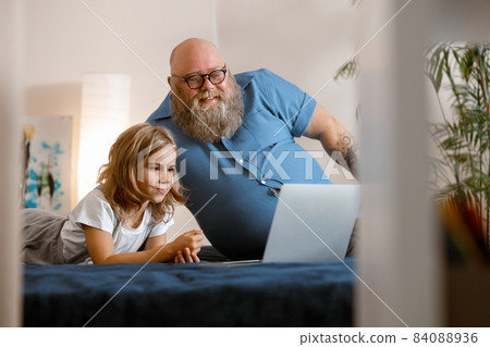 Positive father with beard and little girl take part in video conference via laptop on bed Positive father with beard and little girl take part in video conference via laptop on bed 84088936