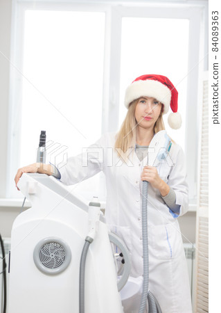 Portrait of a beautiful cosmetologist woman standing in a cosmetology office with a photo rejuvenation tool in her hands and a Santa Claus hat ready to celebrate Christmas and New Year and receive 84089363