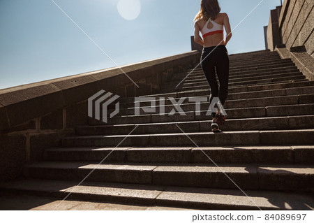 Young sportswoman trains running up stone stairs on sunny summer day Young sportswoman trains running up stone stairs on sunny summer day 84089697