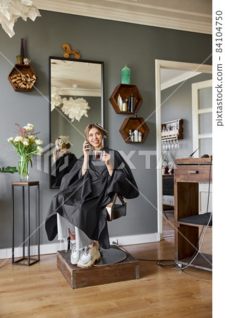 Happy blonde caucasian woman is using smartphone while sitting in chair and waiting in hairdresser's salon 84104750